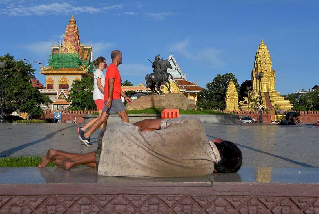 A homeless man lays on the floor as a couple wearing face masks walk past in Phnom Penh on June 23, 2020