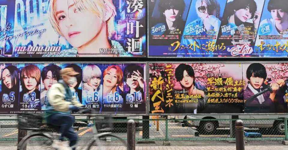 A man rides a bicycle past billboard advertisements for host clubs, establishments that offer entertainment with male companions, along a street in the Kabukicho area of Shinjuku Ward in Tokyo. Host club operators are known for their ties with the criminal underworld.